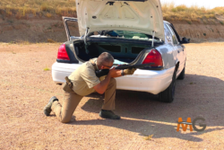 Participant crouching behind car with open trunk with aimed shotgun in shotgun firearms training in Colorado
