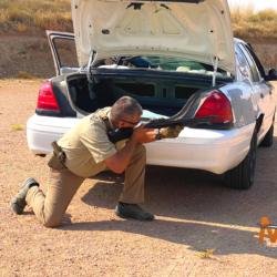 Participant crouching behind car with open trunk with aimed shotgun in shotgun firearms training in Colorado